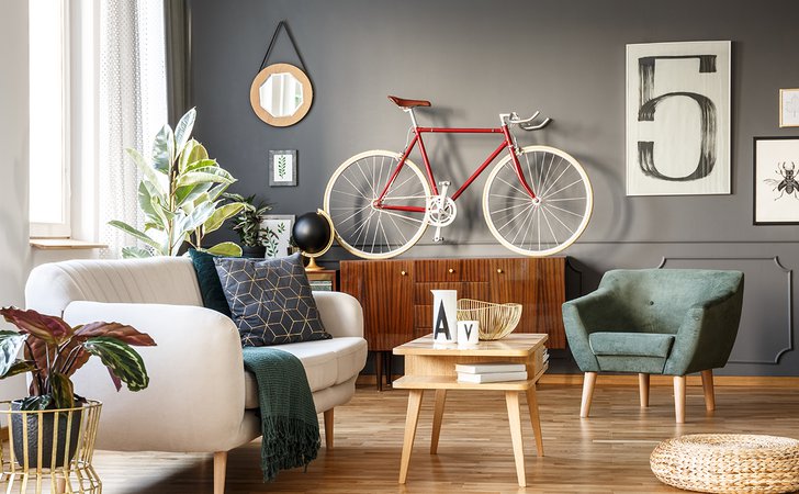 Vintage unique living room interior with good vibes with white comfortable sofa with cushions and blanket and green armchair, cupboard, small table, red bicycle and lots of details including a poster with number five on gray wall